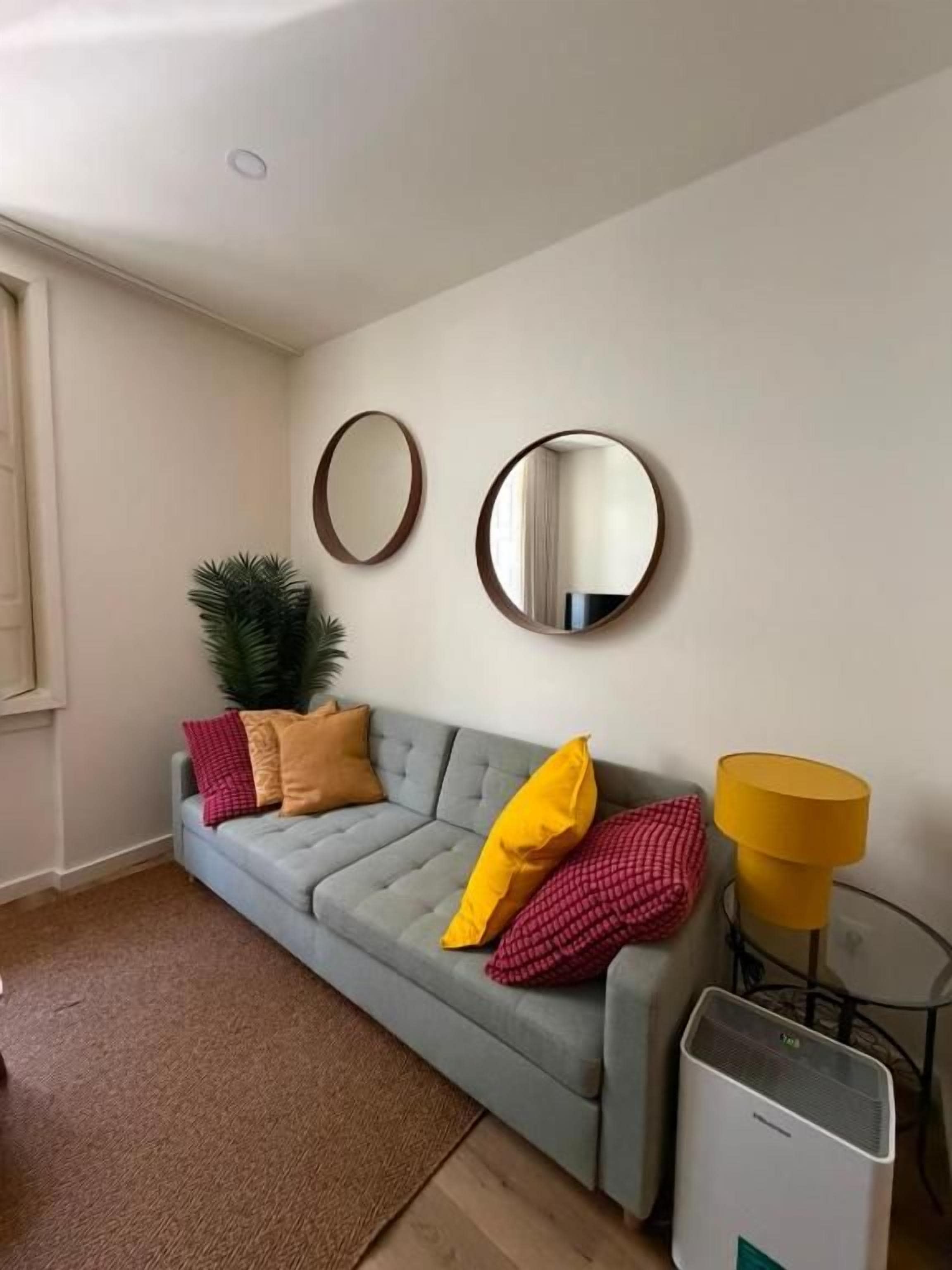 Een woonkamer met een grijze bank, twee ronde spiegels, een gele lamp en een klein tafeltje.