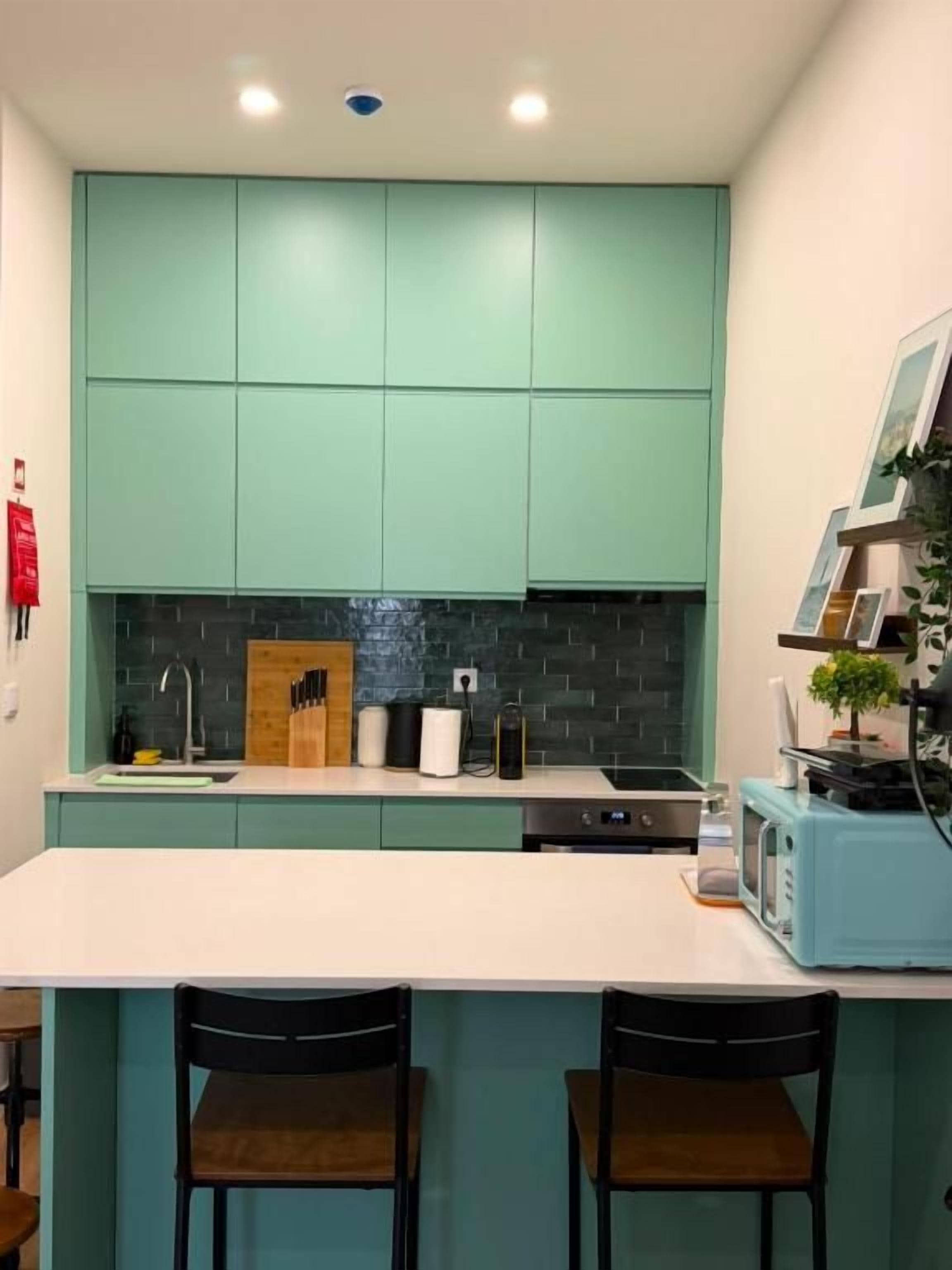 A kitchen with teal cabinets, a white countertop, and two black chairs.