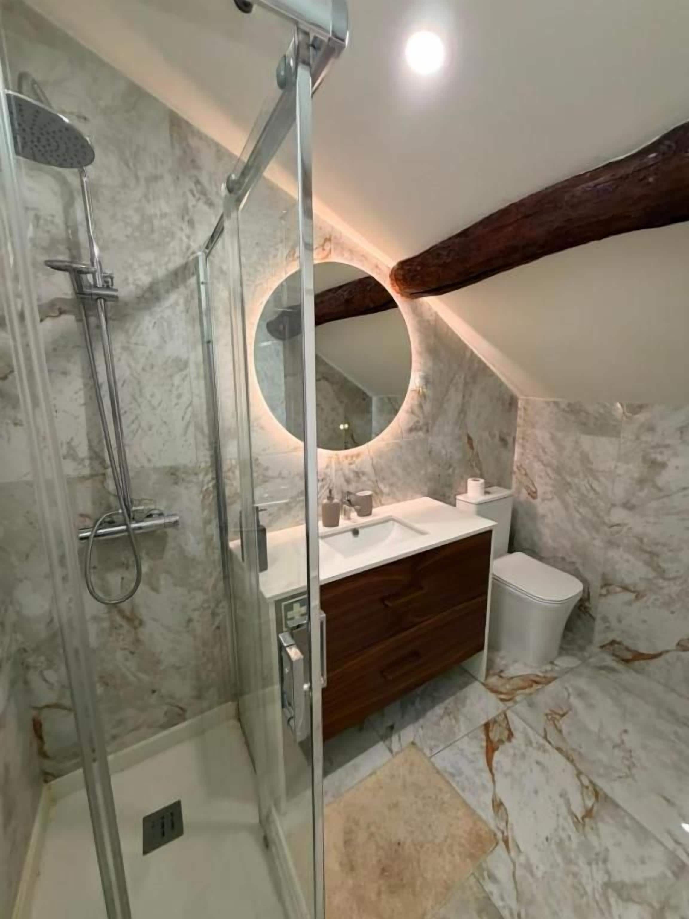 A bathroom with a glass-enclosed shower, a wooden vanity, a round mirror, and marble walls.