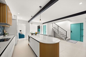 Eine moderne Küche mit einer zentralen Kochinsel, einem Essbereich mit rundem Tisch und einer Treppe zu einer türkisfarbenen Tür.