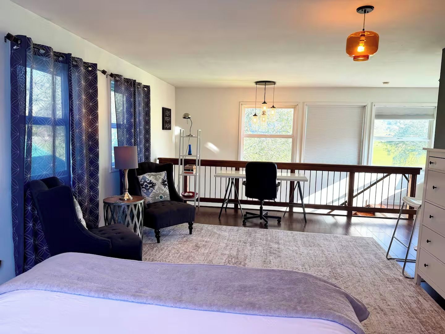 Un dormitorio con cama, escritorio, silla, cómoda y balcón con barandilla.