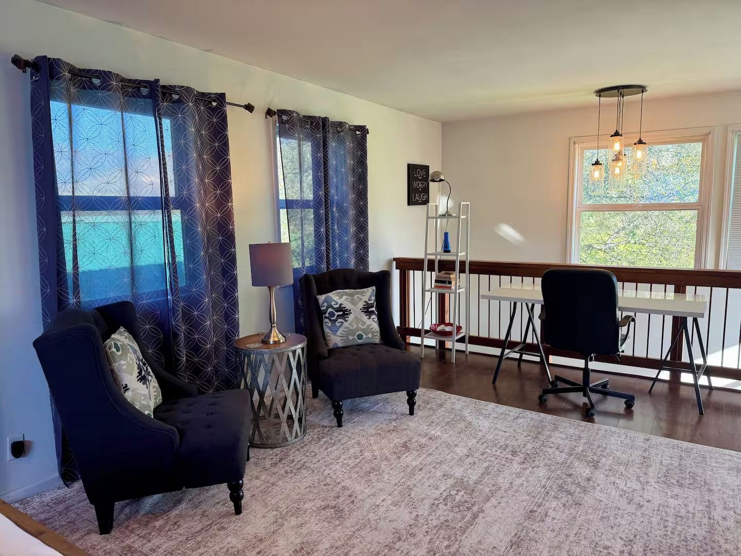 Ein Wohnzimmer mit einer Couch, einem Sessel, einem Beistelltisch, einer Lampe und Blick ins Freie durch große Fenster.