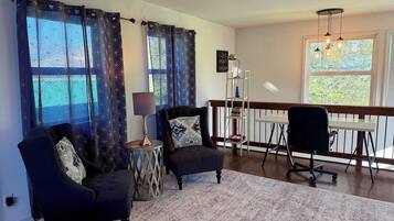 Ein Wohnzimmer mit einer Couch, einem Sessel, einem Beistelltisch, einer Lampe und Blick ins Freie durch große Fenster.