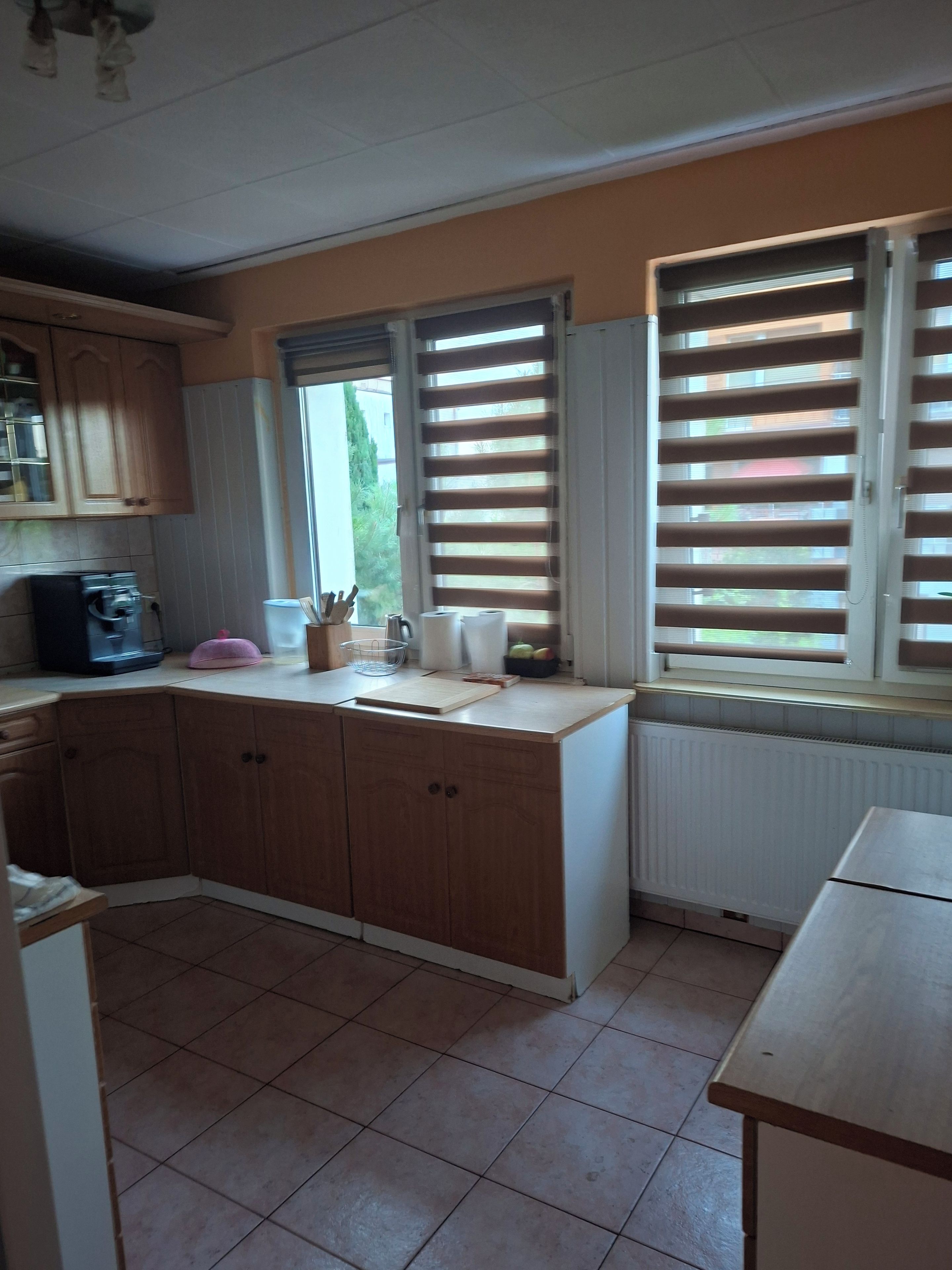 Eine Küche mit Holzschränken, einer Arbeitsplatte, einer Kaffeemaschine und einem Fenster mit Jalousien.
