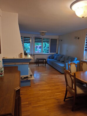 A living room with a blue sofa, wooden dining table, and a kitchen area with a counter. - Spacious Home with fireplace, garden and ponds. (Kołobrzeg)