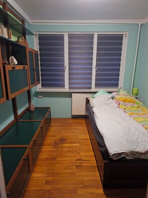 A bedroom with a bed, a window with blinds, a radiator, and a shelf with books and a basket. - Spacious Home with fireplace, garden and ponds. (Kołobrzeg)