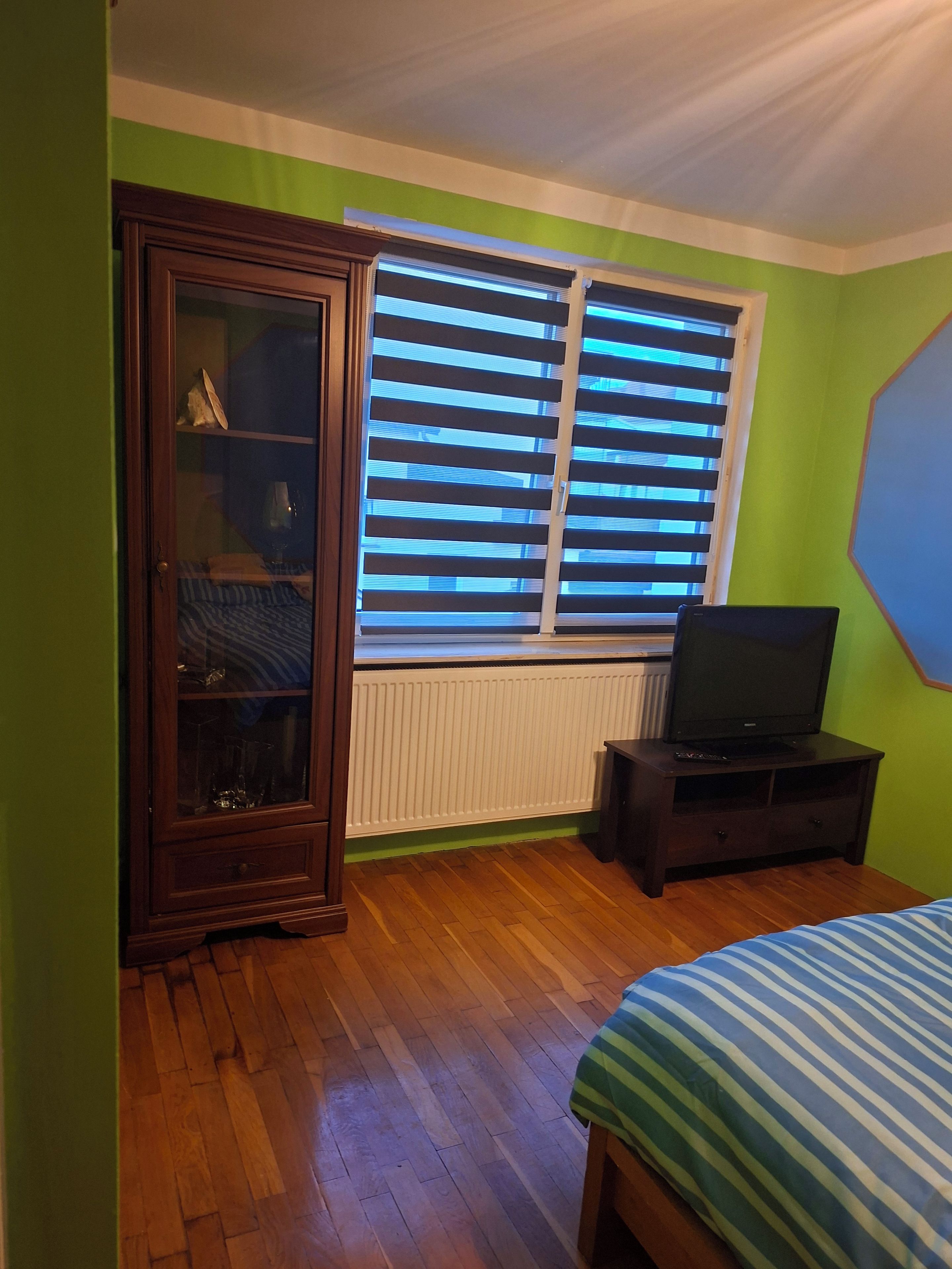 Une pièce avec un meuble en bois, un téléviseur posé sur un meuble, un radiateur et une fenêtre avec des stores.