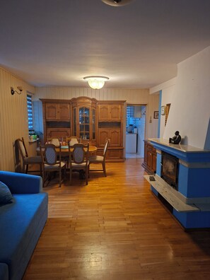 A living room with a blue sofa, wooden dining table, and chairs, a fireplace, and a kitchen in the background. - Spacious Home with fireplace, garden and ponds. (Kołobrzeg)