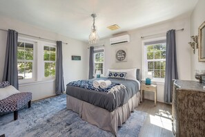 Ein Schlafzimmer mit einem großen Bett, zwei Nachttischen, einer Chandelüster und Blick auf Bäume durch die Fenster.