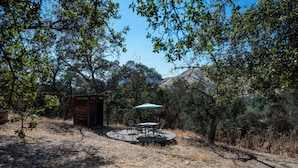 A wooden cabin with a circular deck and a table with chairs, surrounded by trees and dry grass. - Agate Ranch | 2 BD + Views + EV + Walk to River (Three Rivers)