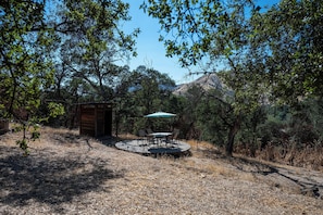 Eine Holzhütte mit einer runden Terrasse und einem Tisch mit Stühlen, umgeben von Bäumen und trockenem Gras.