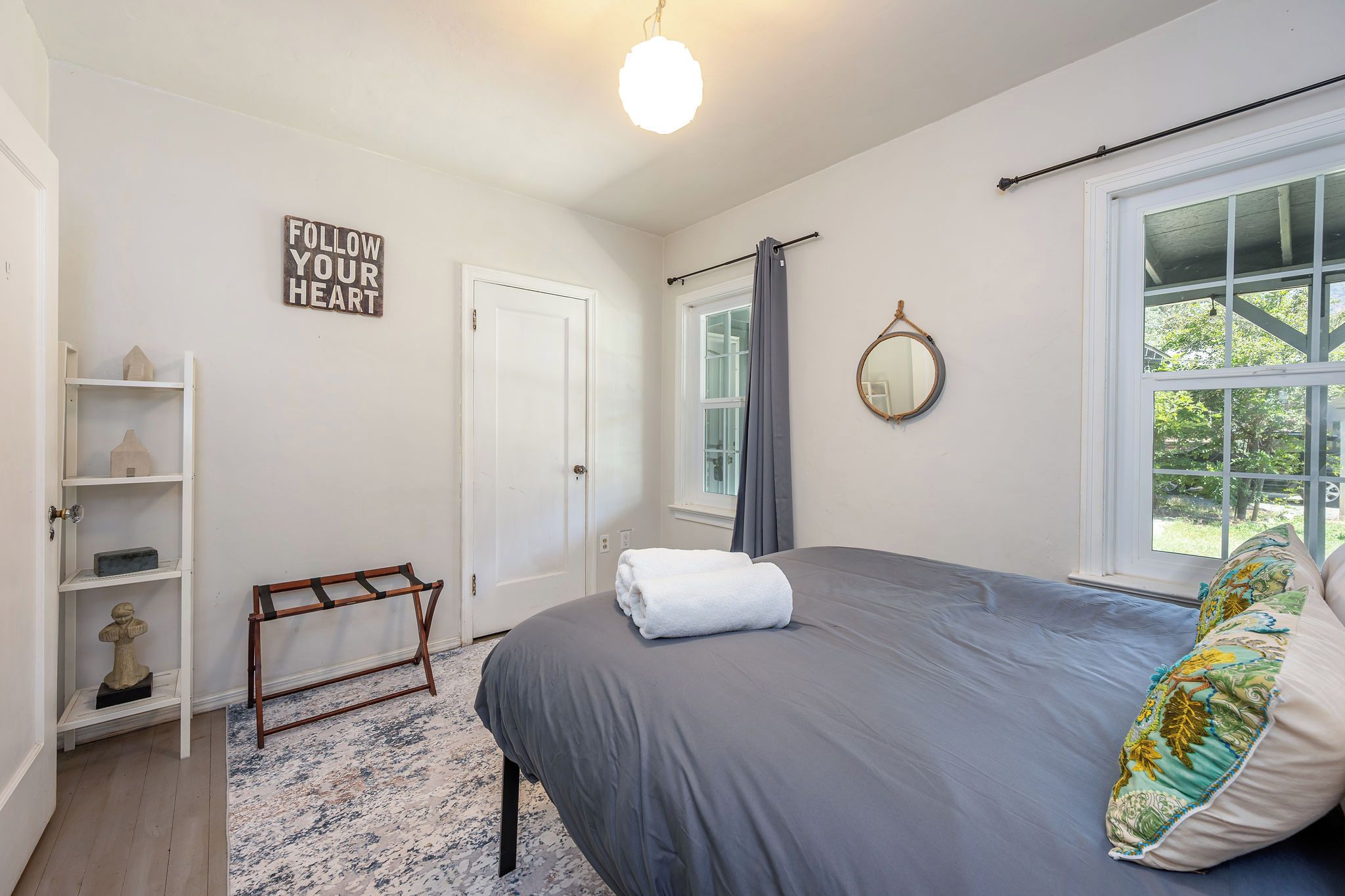 A bedroom with a bed, a bedside table, a mirror, a window with curtains, a door, and a shelf with decorative items.