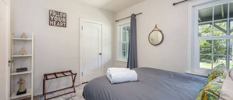 A bedroom with a bed, a bedside table, a mirror, a window with curtains, a door, and a shelf with decorative items.
