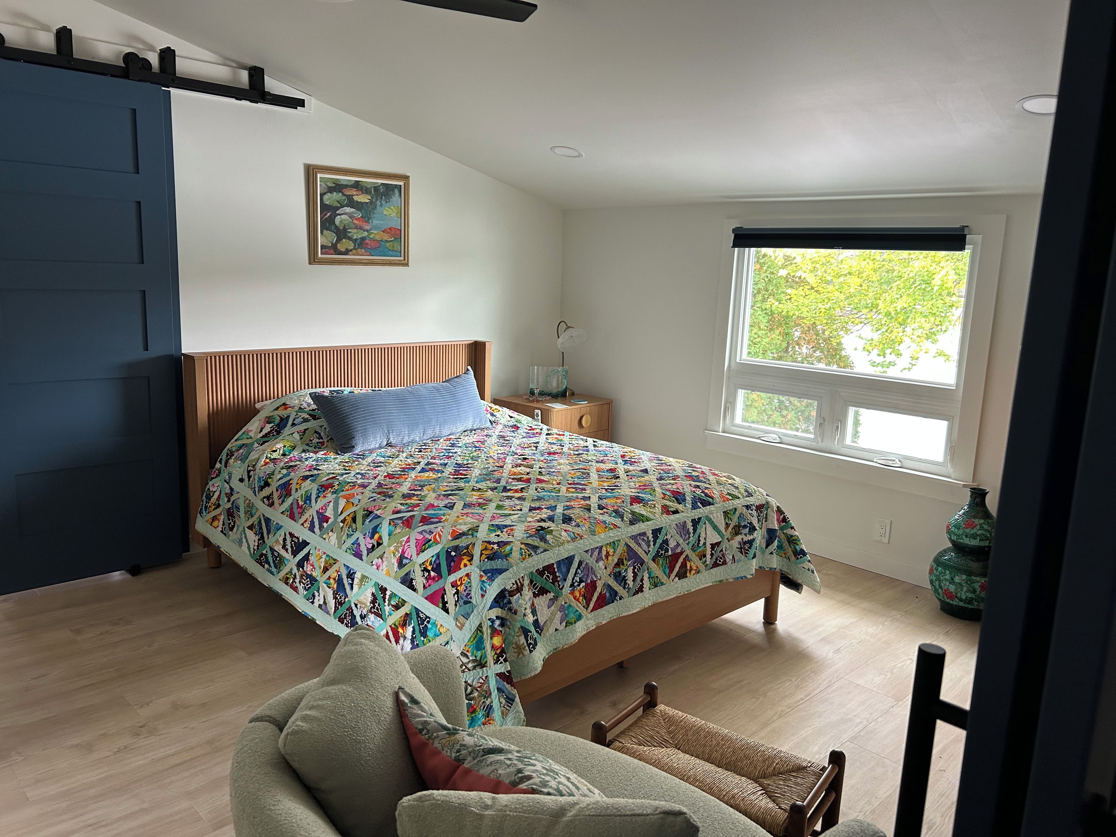 Ein Schlafzimmer mit einem hölzernen Bett, einer farbenfrohen Stegdecke, einem Fenster mit Blick, einer blauen Tür und einem kleinen Tisch mit einer Vase.
