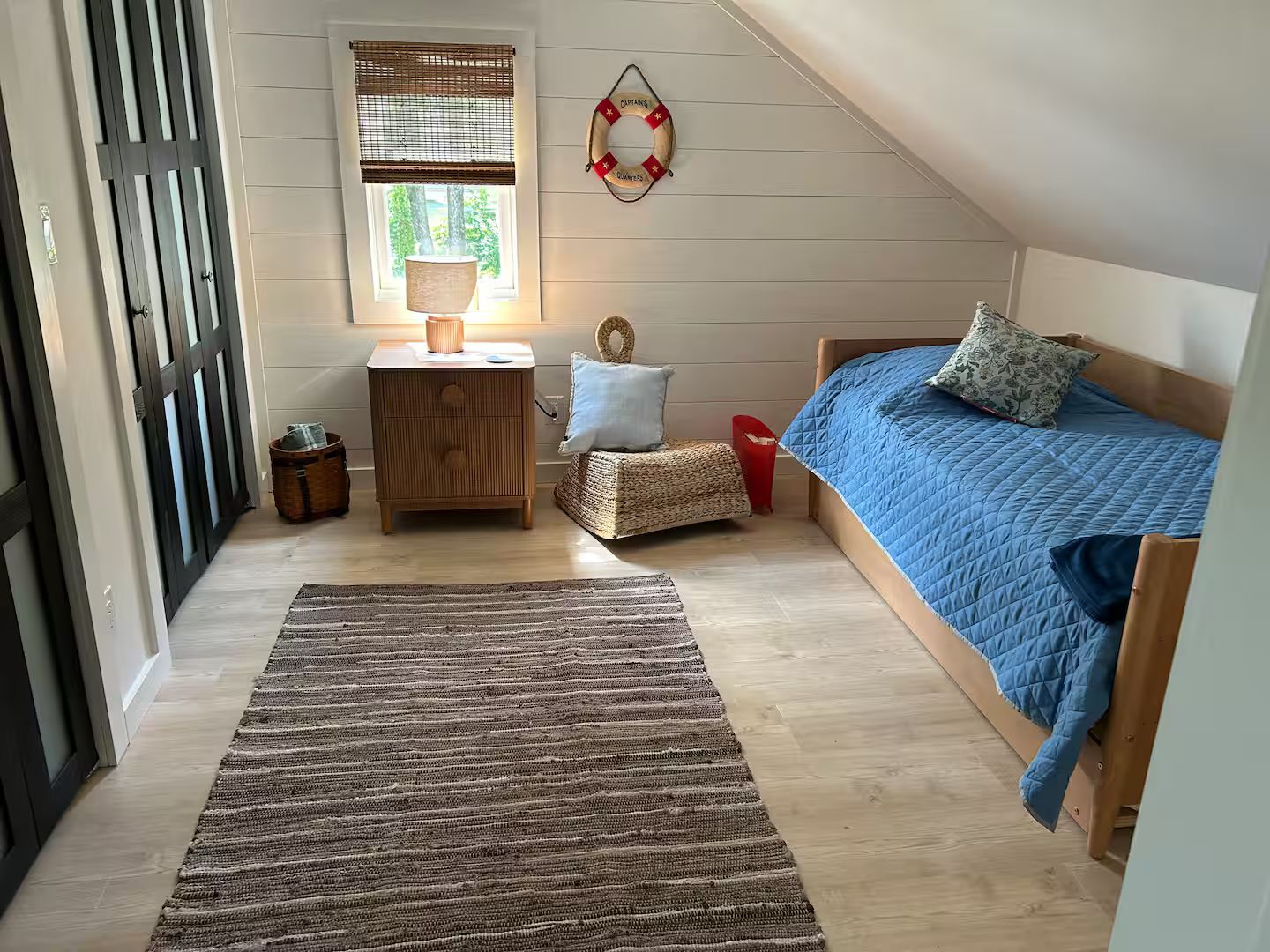 A bedroom with a sloped ceiling, a bed with blue bedding, a bedside table with a lamp, a small stool, and a window with a view of trees.