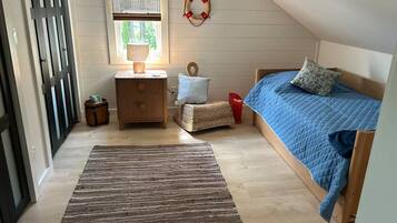 A bedroom with a sloped ceiling, a bed with blue bedding, a bedside table with a lamp, a small stool, and a window with a view of trees.