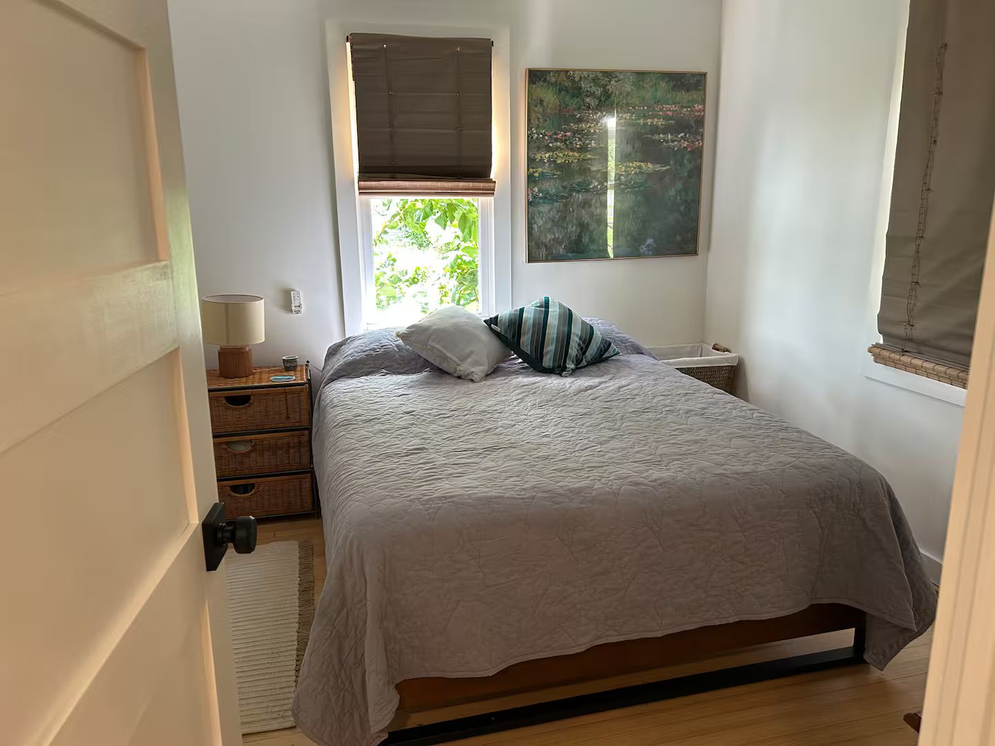A bedroom with a bed, a nightstand, a lamp, a window with a view of greenery, and a painting on the wall.