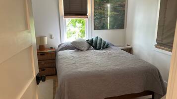 A bedroom with a bed, a nightstand, a lamp, a window with a view of greenery, and a painting on the wall.