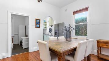 A dining area with a wooden table and white chairs, a chandelier, and a window with a view of greenery.