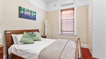A bedroom with a bed, a nightstand, a wall-mounted air conditioner, a window with blinds, and a door.