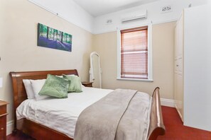 Un dormitorio con cama, mesita de noche, aire acondicionado de pared, ventana con persianas y puerta.