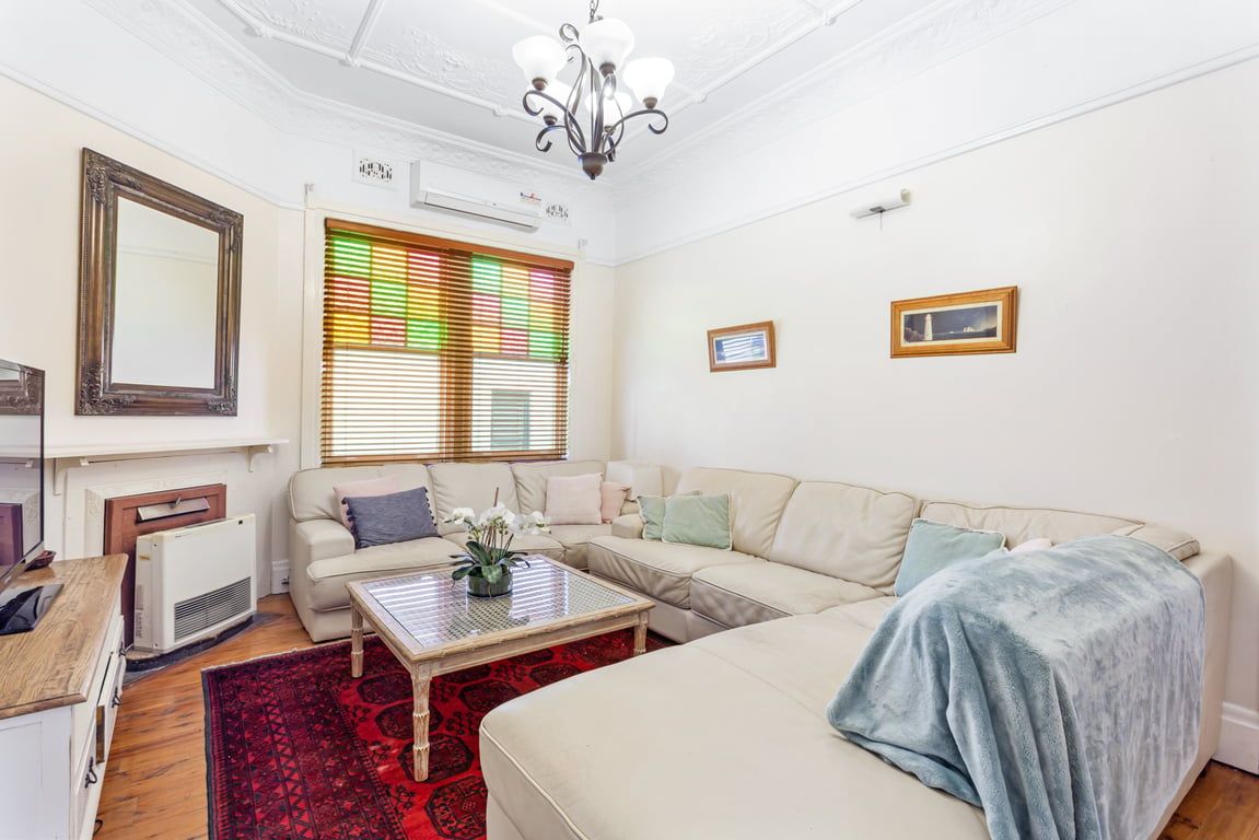 A living room with a sofa, a coffee table, and a radiator. There is a window with blinds and a chandelier.