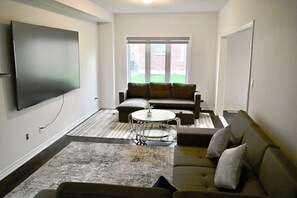 A living room with a flat-screen TV, a sectional sofa, a round coffee table, and a patterned rug. - Luxury 4BR Detached Home 4 Beds plus 2 Sofa Bed Sleeps 10 Close to Niagara Falls (Thorold)