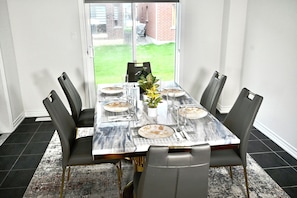 A dining table set for six with chairs, a potted plant, and a view of a grassy yard through a glass door. - Luxury 4BR Detached Home 4 Beds plus 2 Sofa Bed Sleeps 10 Close to Niagara Falls (Thorold)