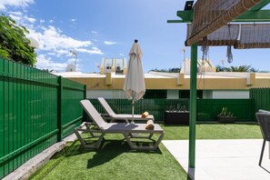 Ein Garten mit Kunstgrasanlage, zwei Liegestühlen, einem kleinen Tisch und einem Pergola mit Dach.