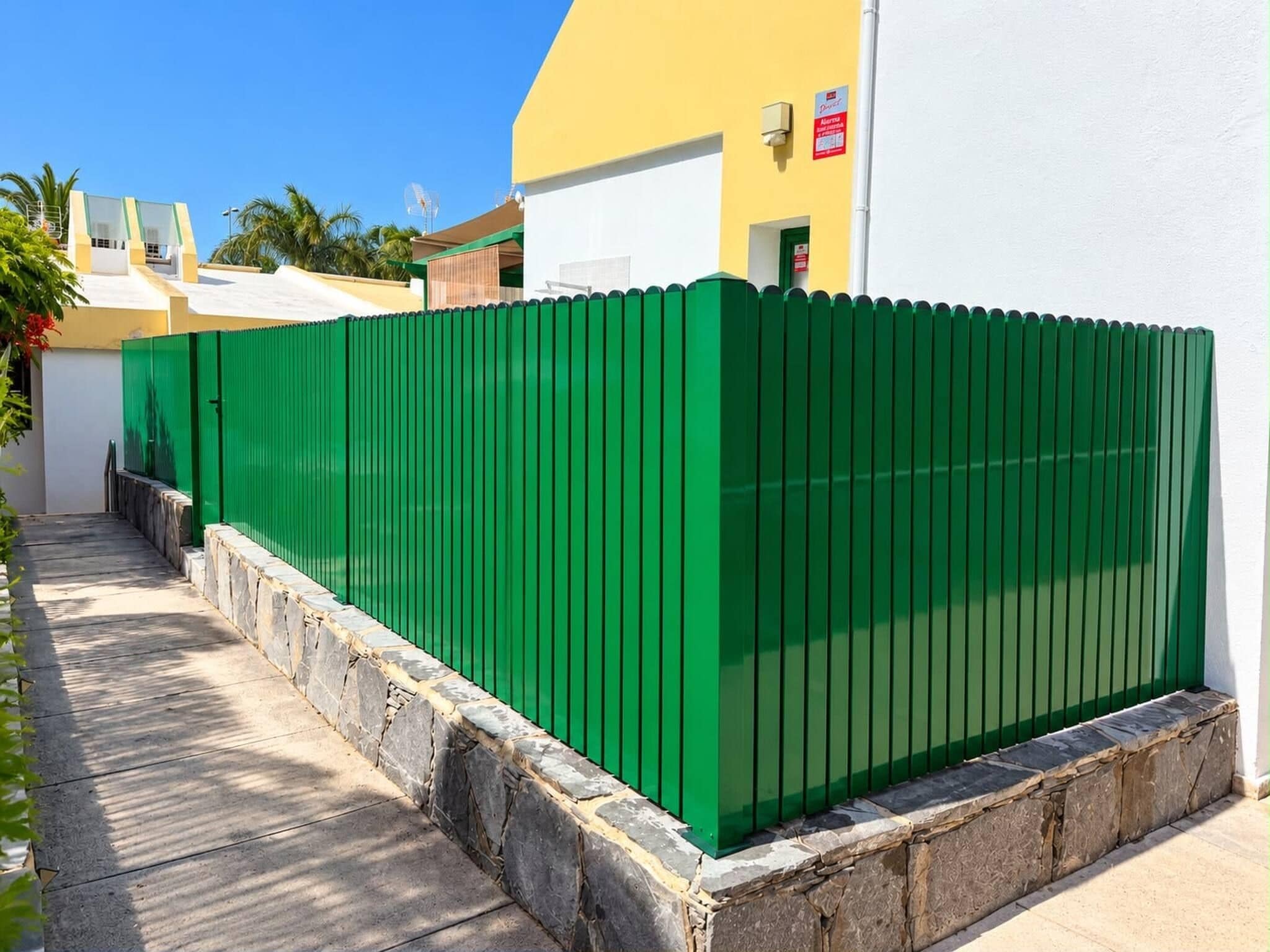 Ein grüner Metallzaun mit steinernen Fundament, neben einem Gebäude mit gelber Fassade und weißer Mauer.