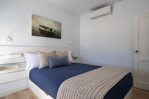 A bedroom with a bed, a nightstand, a wall-mounted air conditioner, and a painting of a boat on the wall. - Maspalomas Bungalow: Private Terrace, Pool, BBQ, and A/C (San Bartolomé de Tirajana)