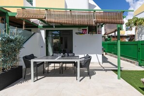 An outdoor patio with a table and chairs, a covered area with a bamboo shade, and a green fence in the background. - Maspalomas Bungalow: Private Terrace, Pool, BBQ, and A/C (San Bartolomé de Tirajana)
