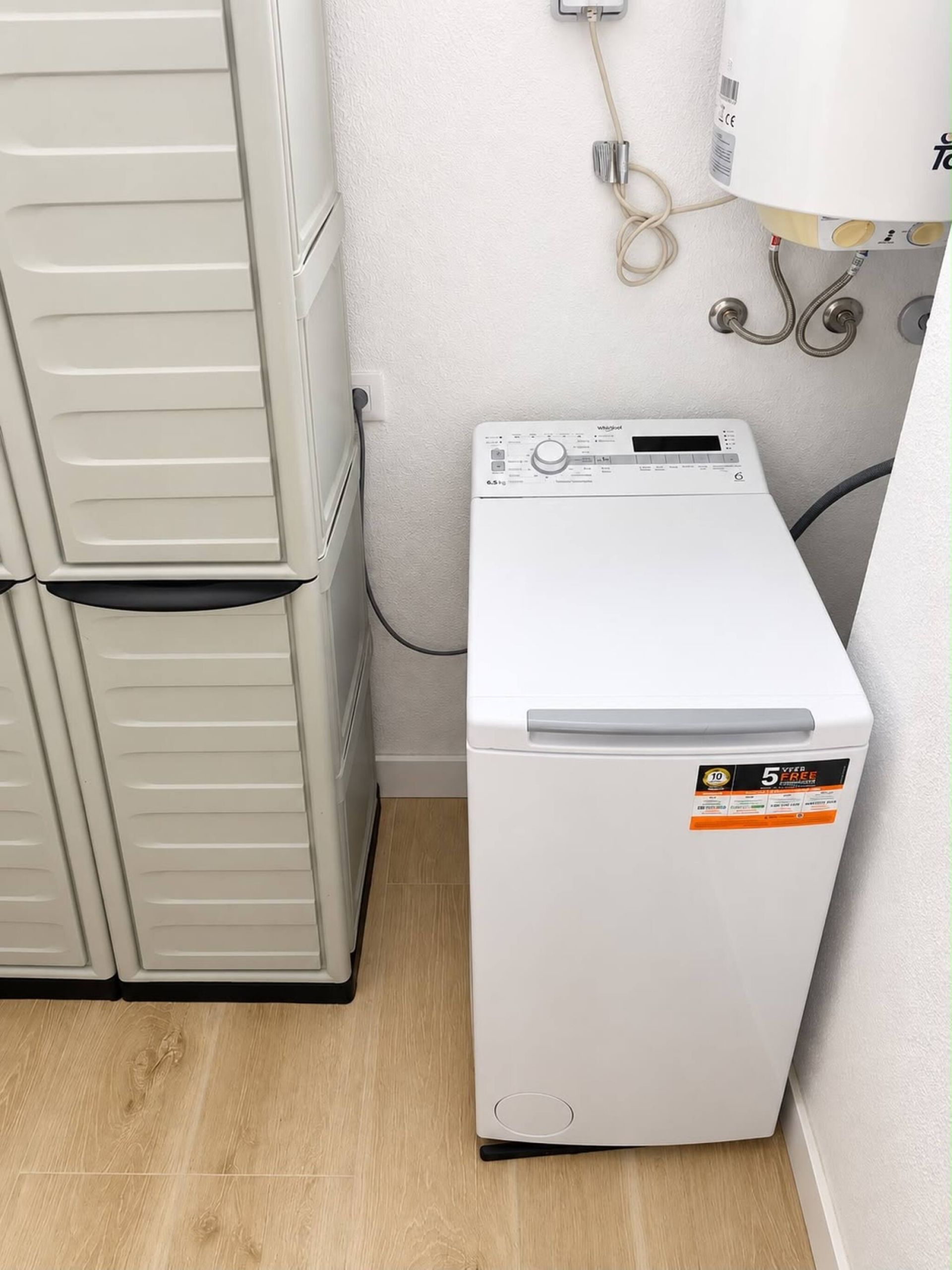 Une petite machine à laver blanche, dotée d’un panneau de commande et d’un autocollant indiquant une garantie de « 5 ans », est placée à côté d’un meuble blanc dans une petite pièce au plancher en bois.