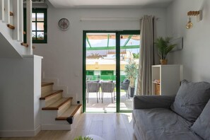 A modern living room with a grey sofa, wooden staircase, and a sliding glass door leading to an outdoor area. - Maspalomas Bungalow: Private terrace, pool, barbecue, and air conditioning (San Bartolomé de Tirajana)