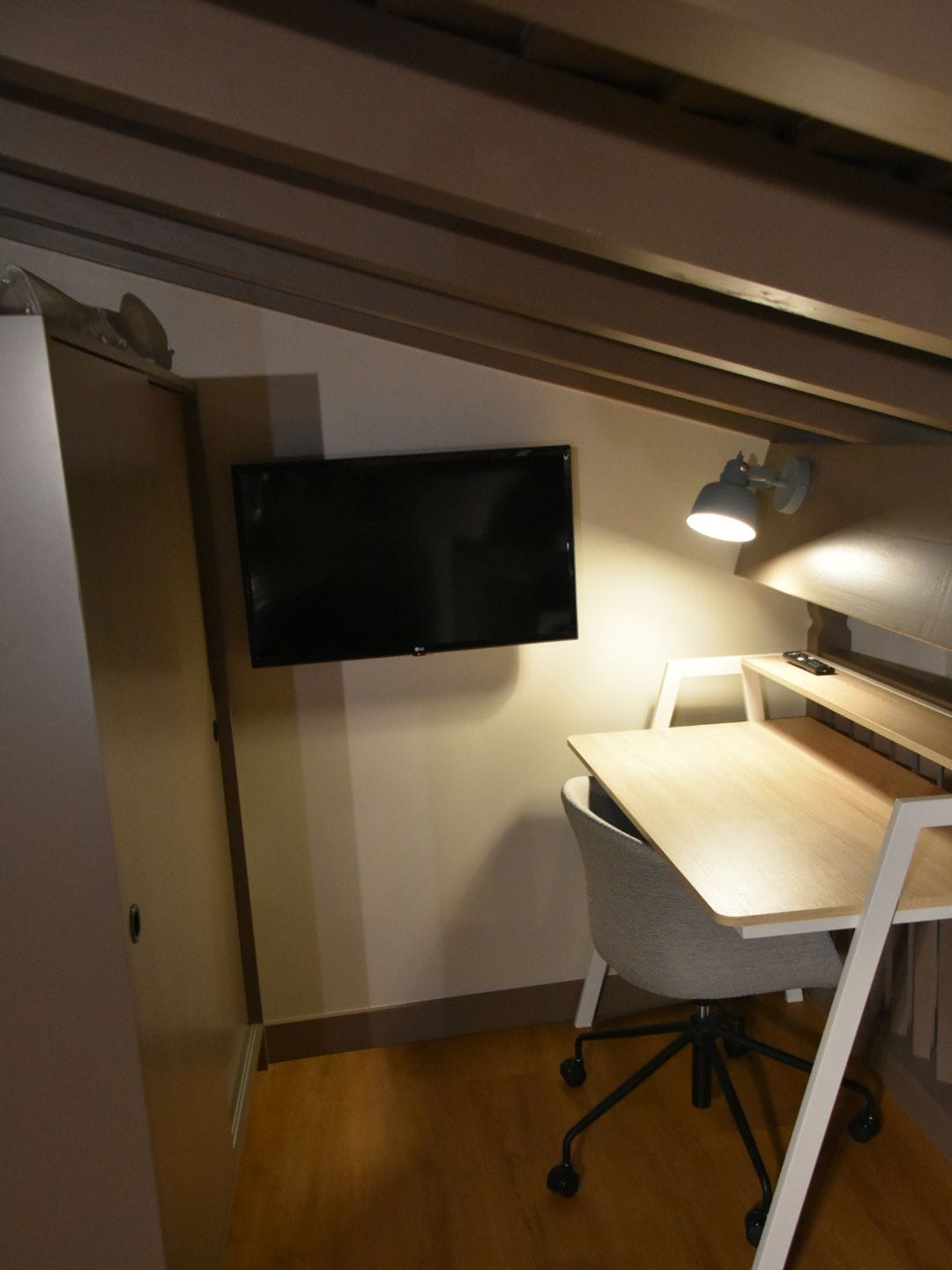 A compact room with a flat-screen TV mounted on the wall, a desk with a chair, and a desk lamp.