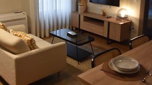 Una sala de estar con un sofá, una mesa de centro, un televisor sobre un mueble y una mesa de comedor para dos personas.