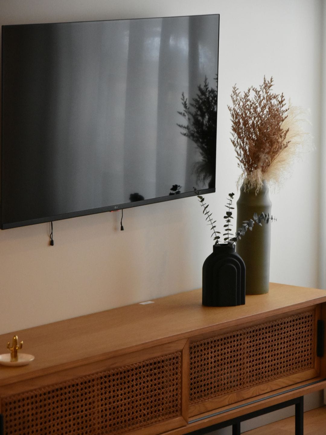 A flat-screen TV mounted on a wall above a wooden console with a cactus and a vase with dried plants.