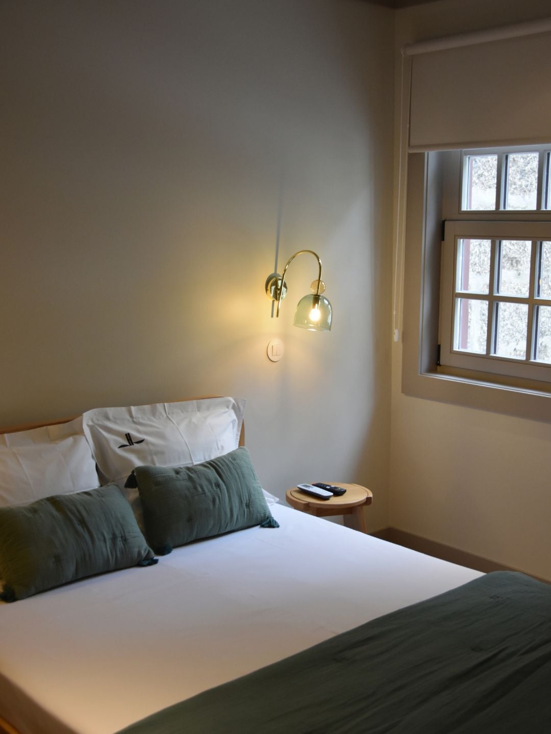 Un dormitorio con cama, almohadas, una mesita de noche con control remoto y una ventana con persianas.