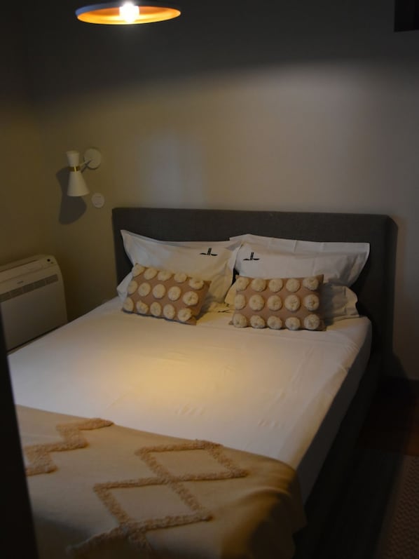 A hotel room with a bed, two pillows with a circular pattern, a wall-mounted lamp, and a heater.
