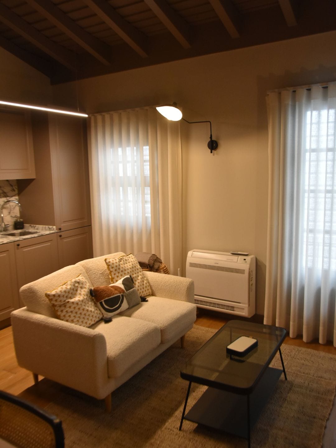 A living room with a sofa, a glass coffee table, a kitchen area with a sink, and an air conditioning unit.