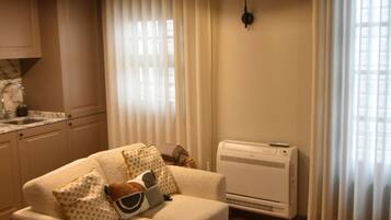 A living room with a sofa, a glass coffee table, a kitchen area with a sink, and an air conditioning unit.