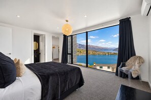 Un dormitorio con un ventanal grande, una cama, una silla y vistas a un cuerpo de agua y montañas.