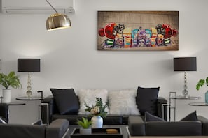 Una sala de estar moderna con un sofá negro, almohadas blancas y esponjosas, una mesa de centro con una planta y un cuadro abstracto colorido en la pared.