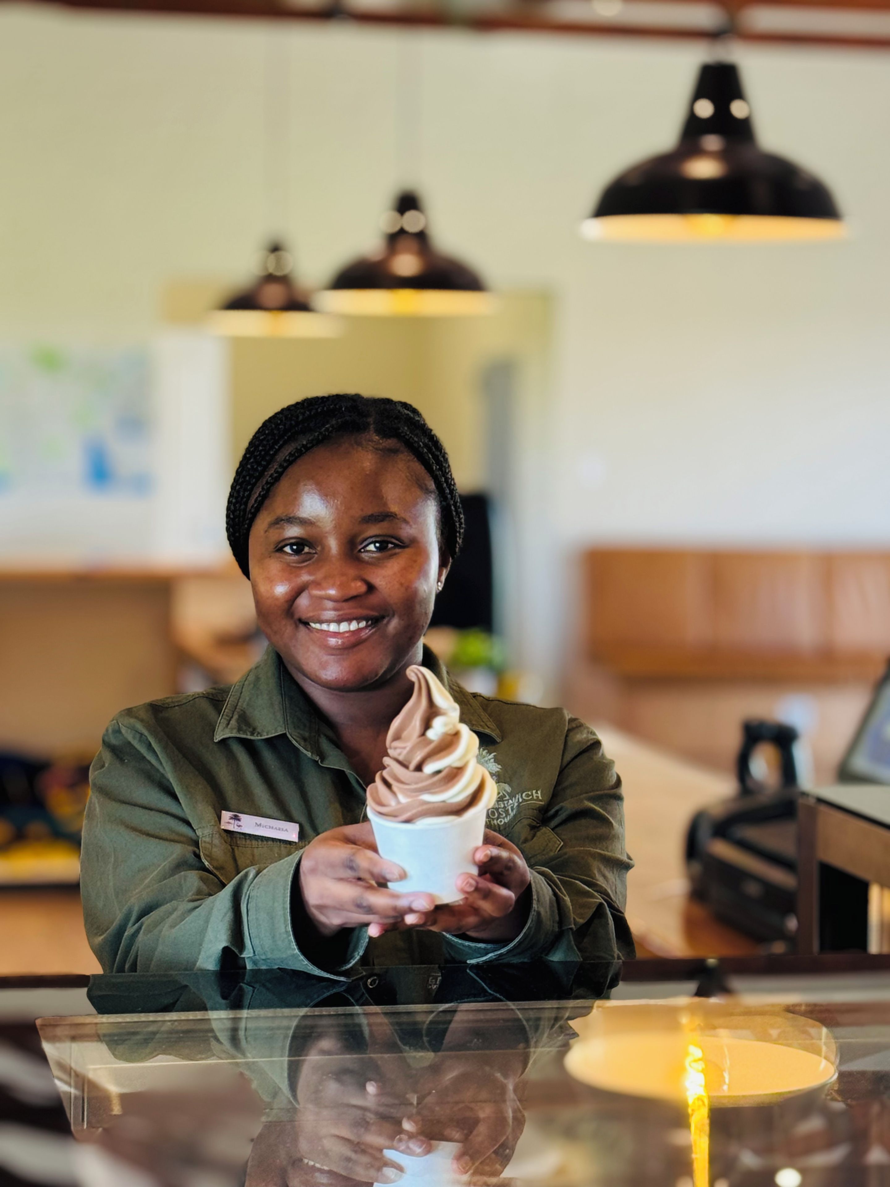 Una persona sosteniendo un vaso de helado en un mostrador, con una placa de identificación, una lámpara y un fondo desenfocado.