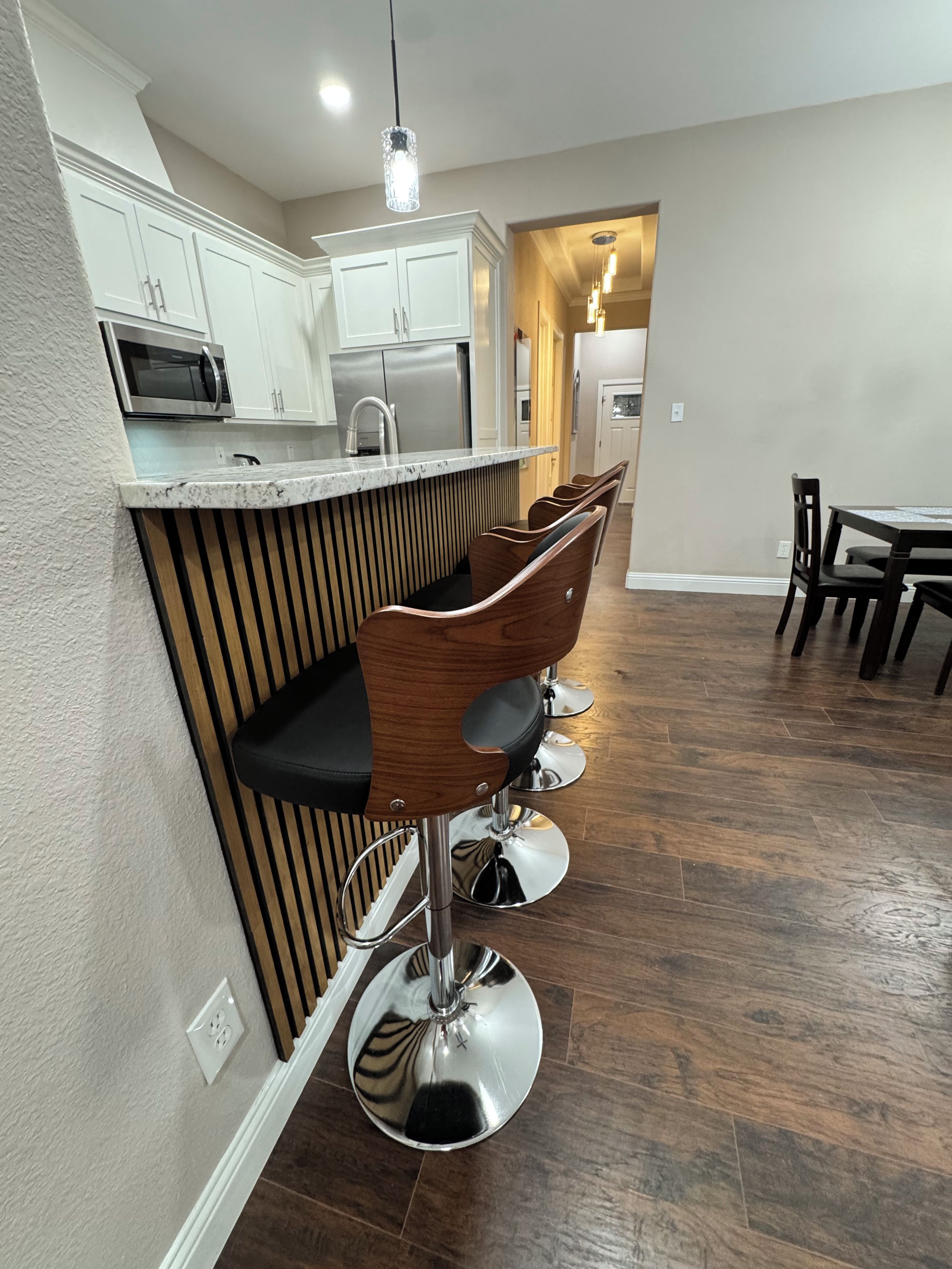 A kitchen with white cabinets, a microwave, and a stainless steel refrigerator. There are wooden bar stools with black seats and chrome bases.