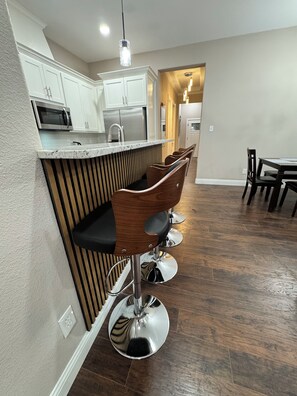 Una cocina con armarios blancos, un microondas y un refrigerador de acero inoxidable. Hay taburetes de bar de madera con asientos negros y bases cromadas.