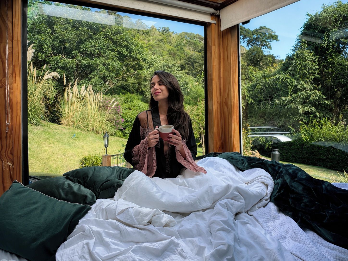 A person sitting on a bed with a view of a lush garden through a large window.