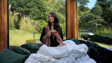 A person sitting on a bed with a view of a lush garden through a large window.