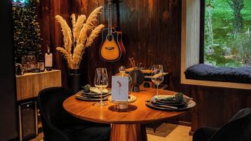 Área de refeições com mesa de madeira, cadeiras, um violão e janela com vista para o verde.
