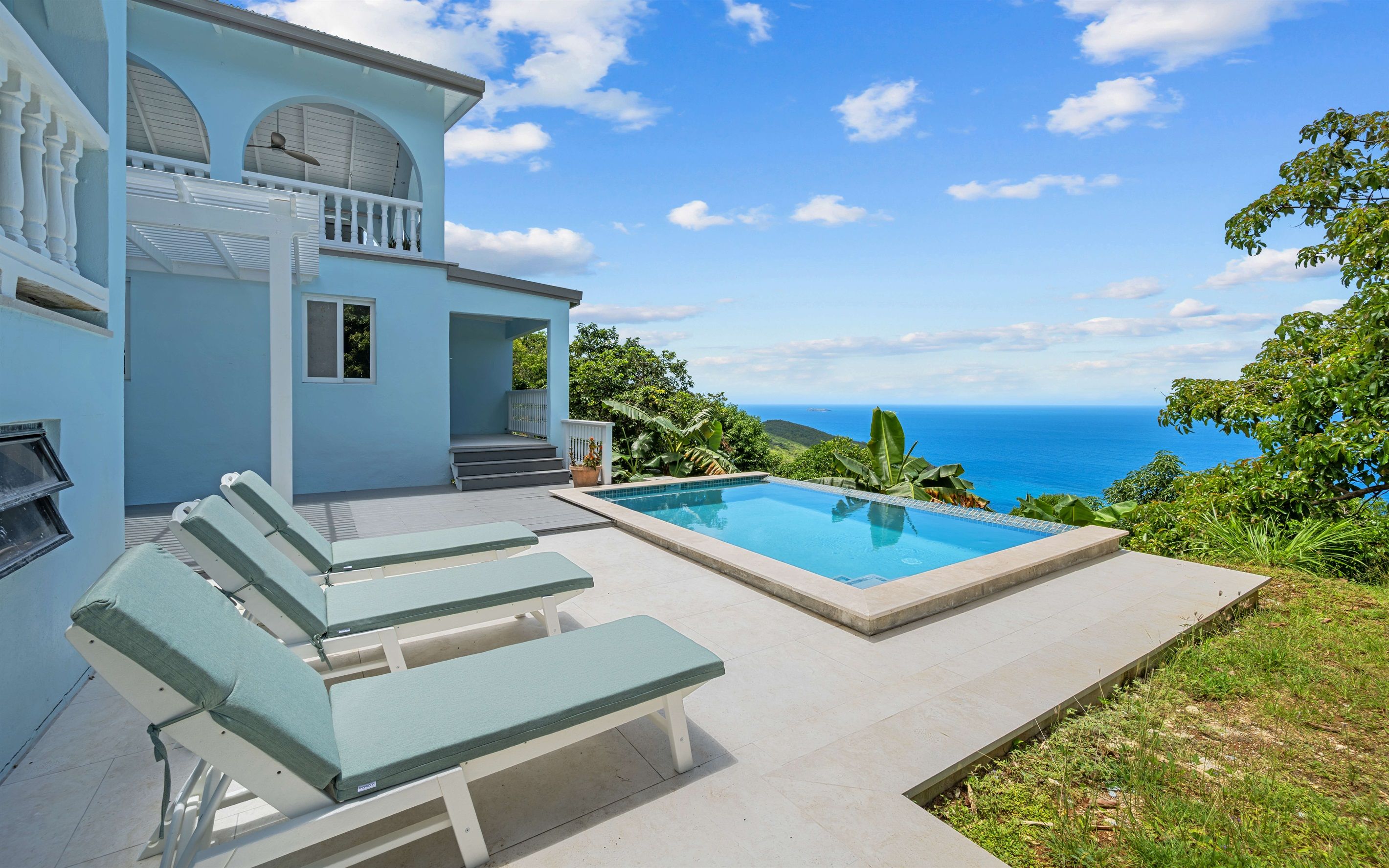 A pool with a view of the ocean, a covered patio with lounge chairs, and a building with a balcony.